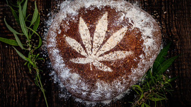 Design on bread shows a leaf pattern. The setting is a wooden surface with herbs and flour around