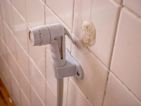 Old worn white plastic bidet hand sprayer mounted on beige ceramic bathroom wall tiles with water hose and rust stains