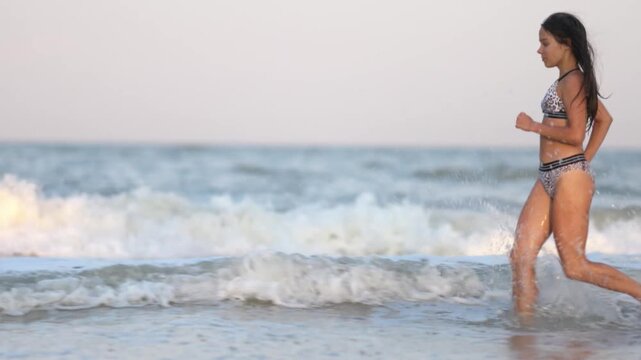 A girl with wet hair in a leopard swimsuit runs along the sea with waves peering into the horizon