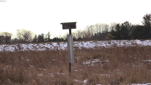 A wide shot of a weathered wooden bluebird nest box marked with number 1358 and fitted with a metal stovepipe predator guard, standing in a dry brown winter field under an overcast sky.