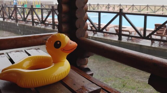 An inflatable duck lies on wooden table in gazebo at recreation center against the backdrop of pool covered with rain