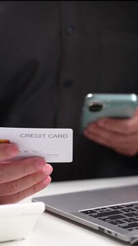 Man wearing black shirt holds plastic credit card, mobile phone near open laptop computer keyboard, making secure digital payment sale inside dark office desk, entering bank data online.