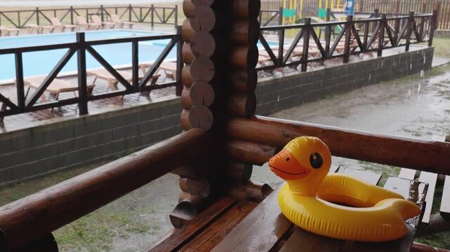An inflatable duck lies on wooden table in gazebo at recreation center against the backdrop of pool covered with rain