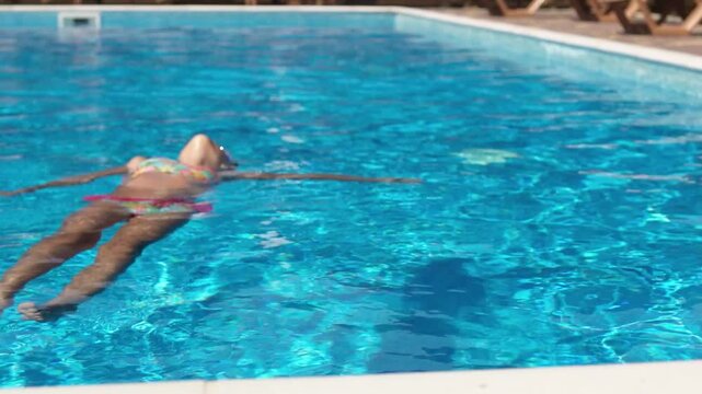 A girl in a pink swimsuit with glasses lies on the water in the pool on her back and enjoys the summer holidays