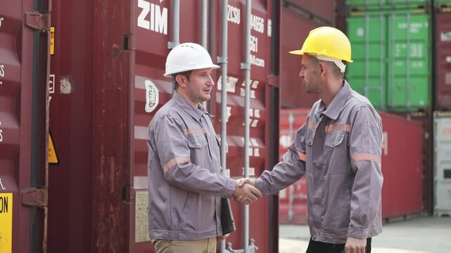 Two male workers in safety helmets and uniforms shake hands in agreement at a sunlit shipping container facility.