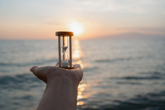 Hand holding hourglass against sunset sea horizon, symbolizing time, mindfulness, patience and slow living. Minimal summer concept with calm ocean background and copy space.