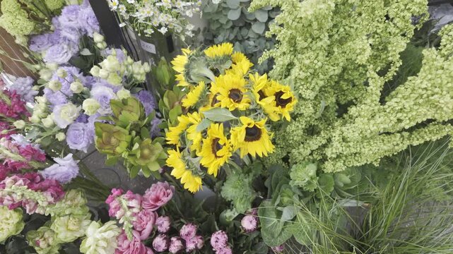 Bright yellow sunflowers and various colorful blossoms create a beautiful floral display at a market. The fresh bouquets and decorative green plants are arranged for sale outdoors.
