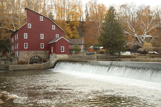 A red wooden mill building with a stone base stands beside a river dam and waterfall in Clinton New Jersey.  A water wheel is visible at the lower wall, with autumn trees in the background.