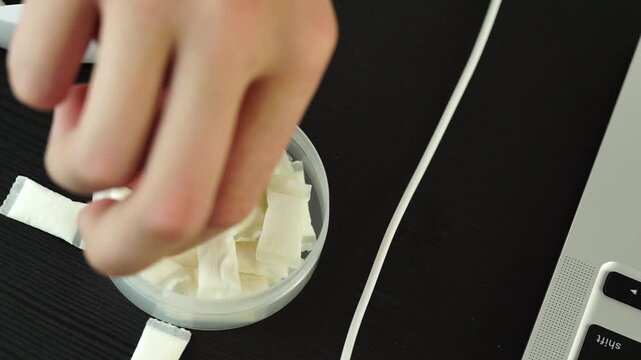 an open round plastic container with white nicotine pouches and snus pads on a dark wooden desk setup with white overhead headphones and a blurry laptop in the background