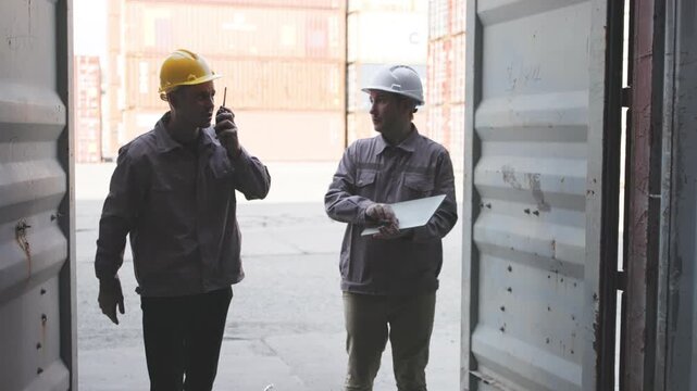 Two male workers in safety helmets examine and discuss a shipping container during operations at a busy logistics terminal.