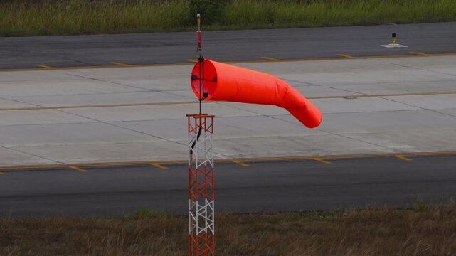 Bright orange windsock blowing in the breeze at an airport. Aviation weather indicator located next to the concrete runway and taxiway lines, providing wind direction and speed for pilots.