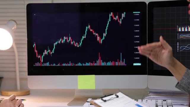Asian businesswoman analyzing financial data and candlestick charts on computer screens. They are discussing investment strategies in a modern office.