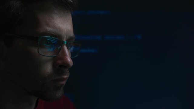 Close up of coder in glasses looks at programming code in a dark, blue script lines reflecting in his lenses. Portrait of software engineer focused on cybersecurity or data science work. Copy space