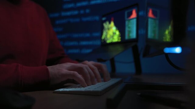 Close-up of a male coder hands typing on a keyboard in a dark high-tech workspace with monitors, data science graphs and programming code projection background, working on a AI-related task.