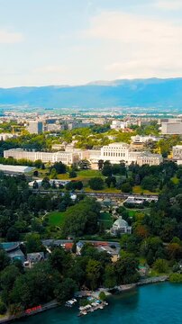Vertical video. Geneva, Switzerland. The Palais des Nations, a vast neoclassical complex of white stone and travertine, overlooks Lake Geneva from the tree-lined Ariana Park under a bright summer sky