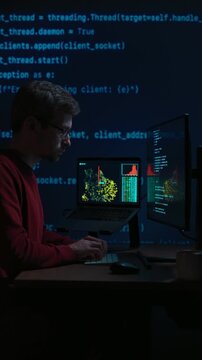 Software engineer in glasses sits at a dark desk with multiple monitors, works on data science or artificial intelligence development. Programmer writing code late at night. Vertical video. Copy space