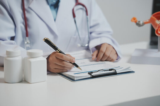 A doctor&rsquo;s hands are shown writing on a medical record form on a clipboard. Two medicine bottles and a stethoscope are visible on the desk.