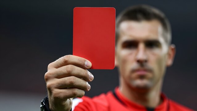 48. [STYLE] of close-up on football referee hand holding up a red card showing penalization with crisp plastic card edges, professional uniform fabric, harsh direct stadium spotlight, vivid solid crim