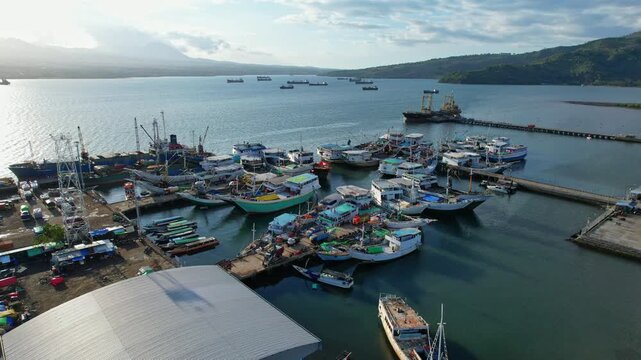 Aerial drone footage of Bima city harbor area at sunset with clouds, in Sumbawa island, Indonesia, with many local big fishing boats at the pier, a big shed, and tanker ships behind in the bay