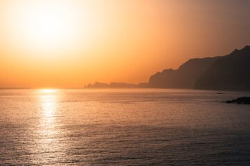Naklejka premium Bright golden sunrise over calm ocean horizon with distant coastal cliffs.