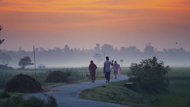 Bogura, Bangladesh - 17 May 2026: View of people walking along a winding rural road amidst misty fields and haystacks under a vibrant orange sunrise sky.