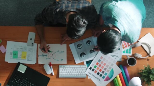 Top down view of smart designer helping manager choosing color from colored palettes. Aerial view of businessman working together and planning together while looking at logo from laptop. Symposium.