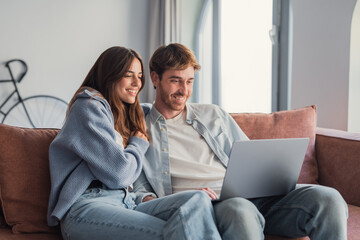 Obraz premium Young couple relaxing on a sofa at home, smiling and sharing a laptop while streaming content together, enjoying cozy, modern domestic leisure and connection