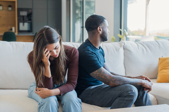 Relationship problems causing sadness and stress for a young adult couple sitting apart on a sofa at home, experiencing emotional distance and disagreement