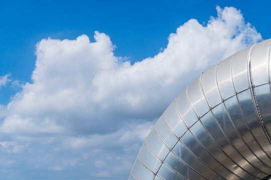 Close-up of a large silver metallic insulation pipe elbow at a petrochemical plant against a bright blue sky with white clouds.