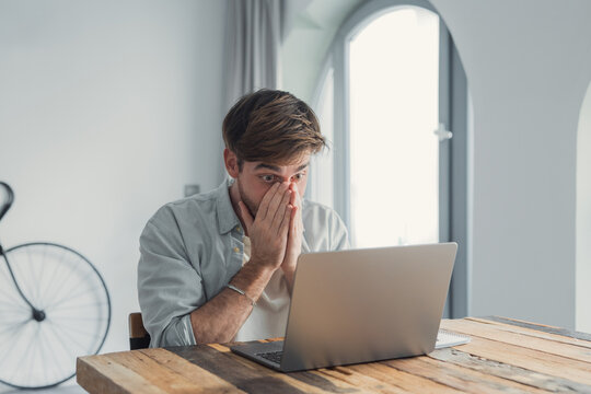 Young man at a wooden desk in a home office, covering his face in shock and stress while staring at his laptop screen, overwhelmed by bad news or a work crisis