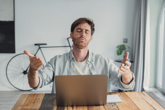 Frustrated young adult man sitting at a wooden table with a laptop, expressing stress and exasperation with eyes closed and hands in a gestural shrug during remote work