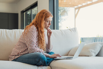 Fototapeta premium Redhead woman sitting on a comfortable sofa, concentrating on her laptop while working from home in a modern living room, representing concepts of freelance work and remote business connectivity