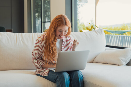 Young redheaded woman on a white couch celebrates good news while working on her laptop at home, smiling with relaxed, casual style in bright natural light