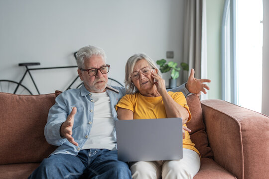 Senior couple sitting on a sofa, looking frustrated and confused while trying to use a laptop at home, experiencing problems and needing help with modern technology