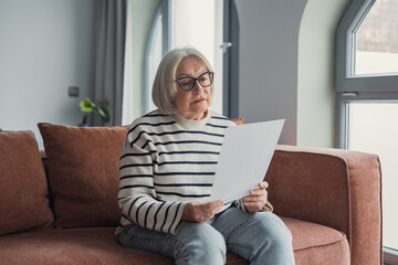 Obraz premium Senior woman wearing glasses and a striped sweater, sitting on a couch in her living room, carefully reading official papers and managing her finances