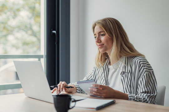 Young blonde woman teleworking from a modern home office, typing on a laptop and jotting notes in a spiral notebook, focused and productive with a coffee nearby