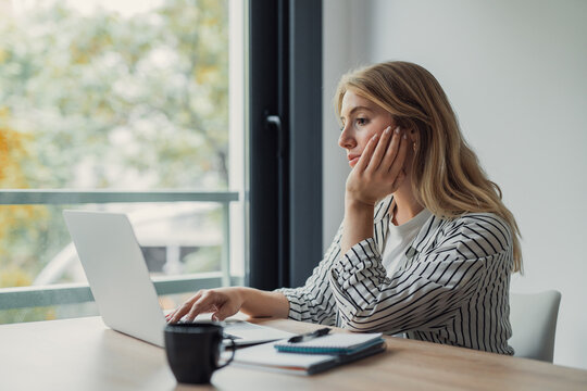 Young woman at a home office desk looking away thoughtfully, distracted and unmotivated while working on a laptop, daydreaming and procrastinating during remote work