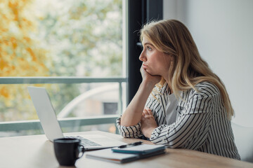 Obraz premium Young woman in a striped shirt sits at a desk with laptop and coffee, daydreaming and staring out a large window during a distracted, unmotivated work-from-home moment