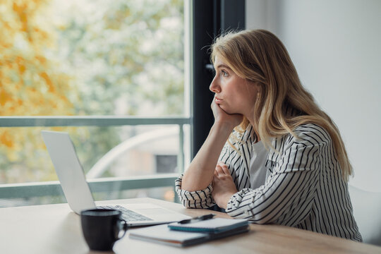 Young woman in a striped shirt sits at a desk with laptop and coffee, daydreaming and staring out a large window during a distracted, unmotivated work-from-home moment