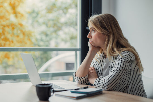 Young adult woman with blonde hair wearing a striped shirt sitting at a table with a laptop and coffee cup, looking out the window, appearing bored or lost in thought