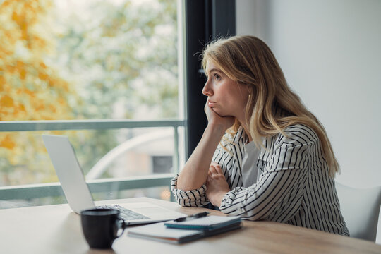 Young woman sitting at a desk with a laptop and coffee, looking out the window while procrastinating and feeling bored or unmotivated during her workday