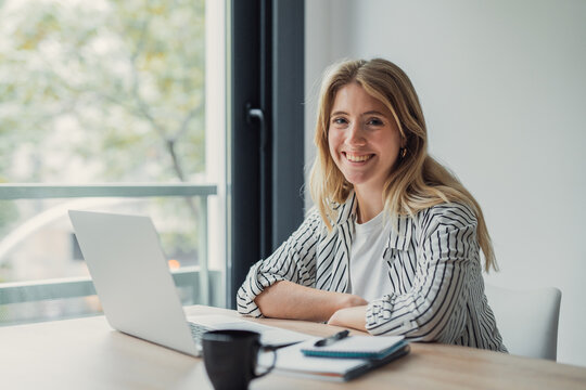 Young woman smiling confidently at her desk with laptop, notebook, pen and coffee, embodying productive remote work lifestyle, modern freelance professional in home office
