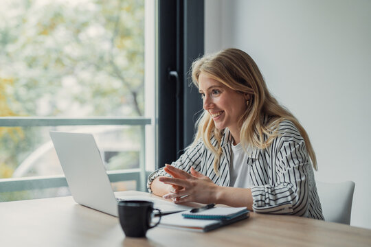 Young adult woman smiling and gesturing during a video call on her laptop, working remotely from her home office with a coffee cup and notebook on the desk