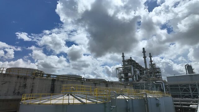 Industrial time-lapse of a large petrochemical refinery plant and storage tanks under a moving cloudy sky.