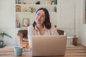 Young professional woman in glasses sitting at a wooden table with laptop and coffee, smiling and gazing away while dreaming and planning during relaxed remote work at home © Daniel