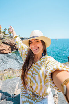 Young smiling woman wearing a hat and striped shirt, taking a selfie while enjoying a sunny day at the coastline, feeling happy and free during her travel
