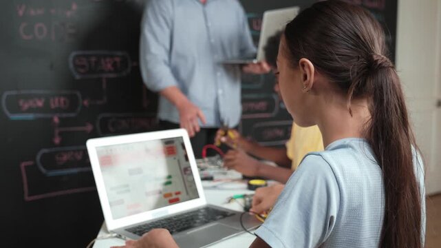 Caucasian girl programming or coding system while african boy fixing model. Teacher holding laptop while looking at diverse children writing web develop program. Innovation education. Edification