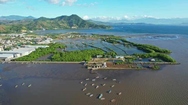 Aerial drone footage of Bima city harbor area, with large green pounds for fish and salt, at sunset with clouds, in Sumbawa island, Indonesia, and mountains in the background, and wide bay