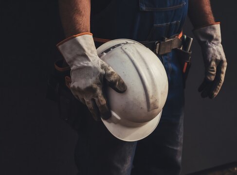 Construction worker holds white hard hat and dirty work gloves. Man wears blue overalls and tool belt. Safety gear displayed against dark background. Labor profession close up.
