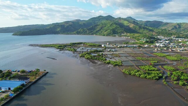 Aerial drone footage of Bima city harbor area, with green pounds for fish and salt, at sunset with clouds, in Sumbawa island, Indonesia, and mountains in the background